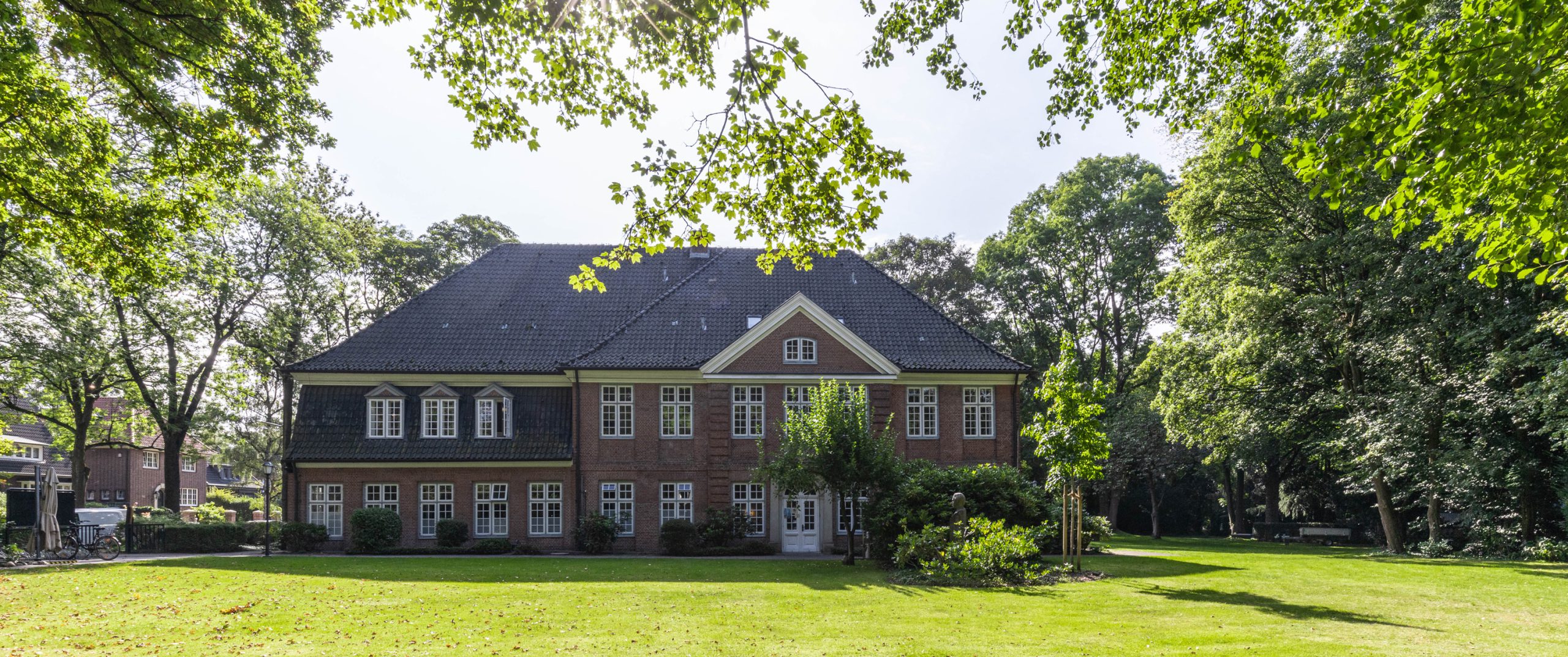 Backsteingebäude mit vielen Fenstern im Grünen, umgeben von Bäumen und Rasen.