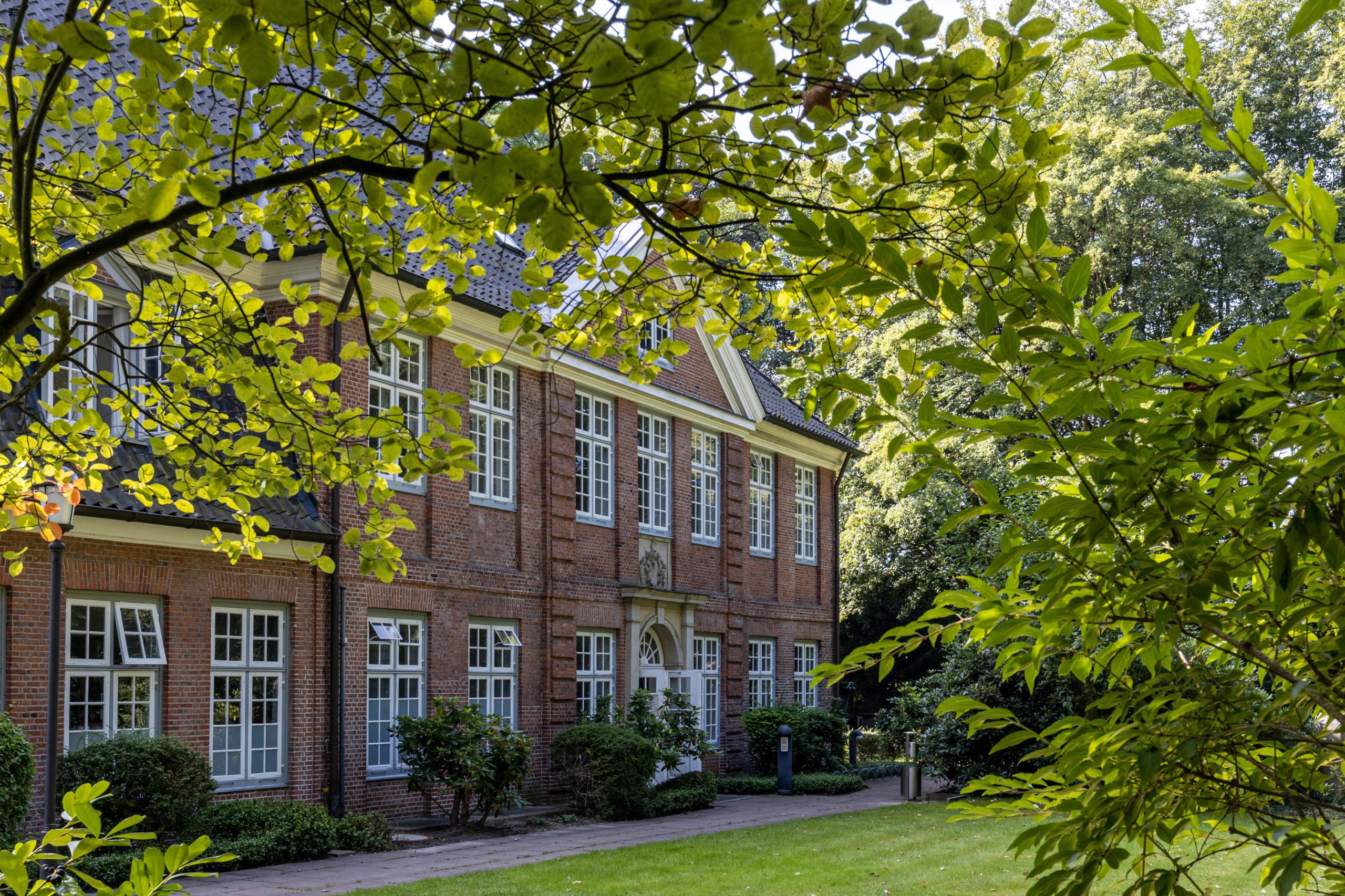 Backstein-Gebäude mit vielen Fenstern, umgeben von grünen Bäumen und Sträuchern.