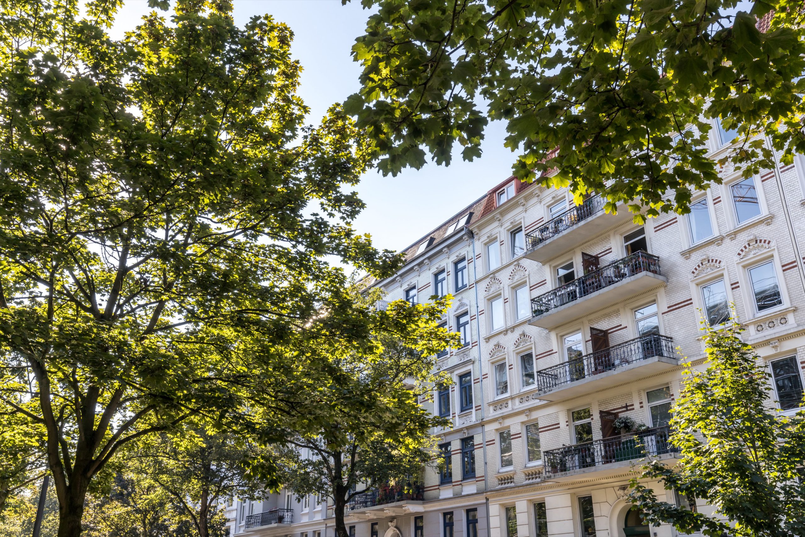 Altbauwohnhaus mit Balkonen hinter grünen Bäumen im Sonnenschein.