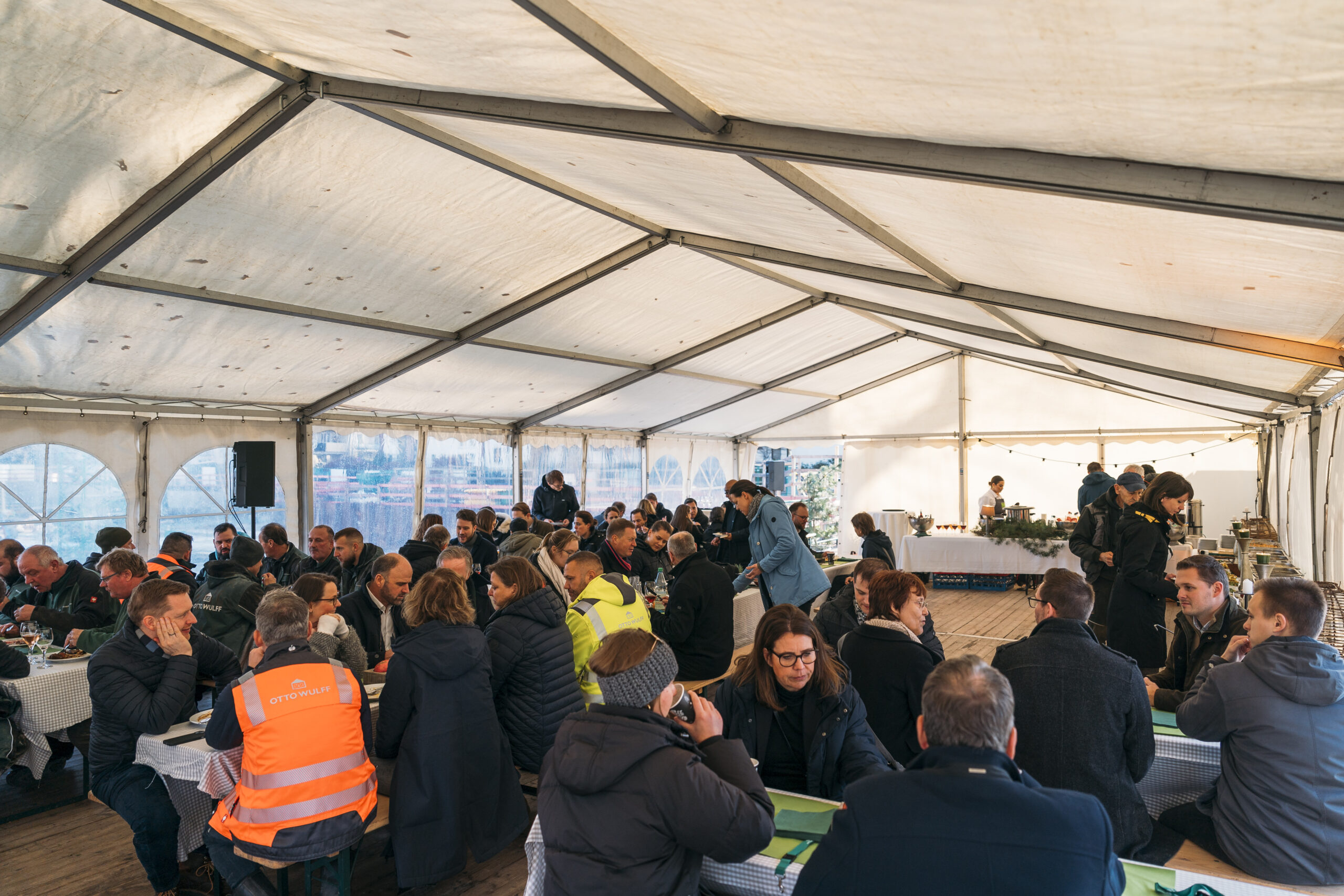 Viele Menschen sitzen in einem Zelt an langen Tischen und essen gemeinsam.
