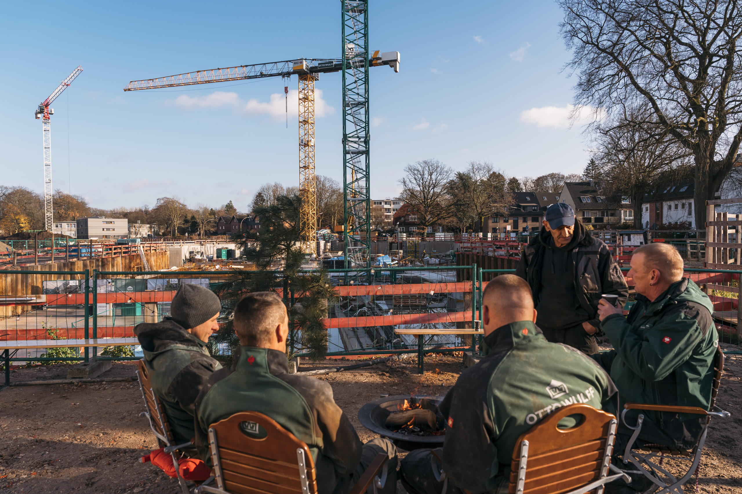 Vier Arbeiter machen Pause vor einer Baustelle mit Kränen im Hintergrund.