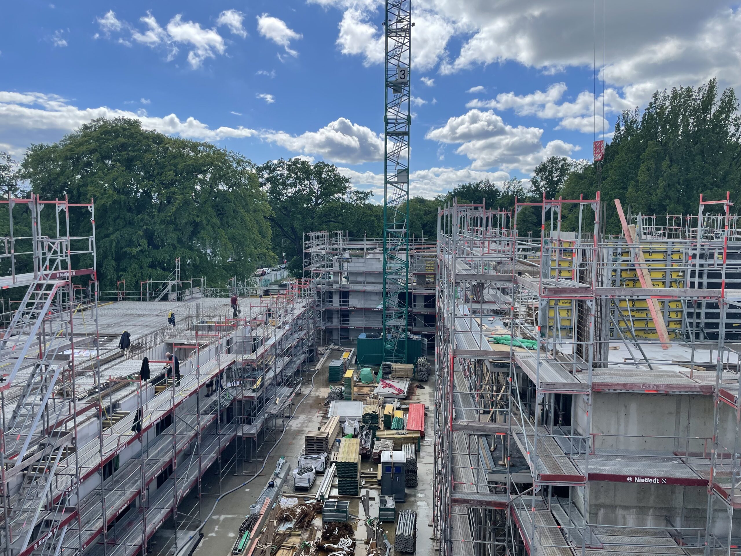Baustelle mit Gerüsten, Baumaterialien und Kran, umgeben von Bäumen.