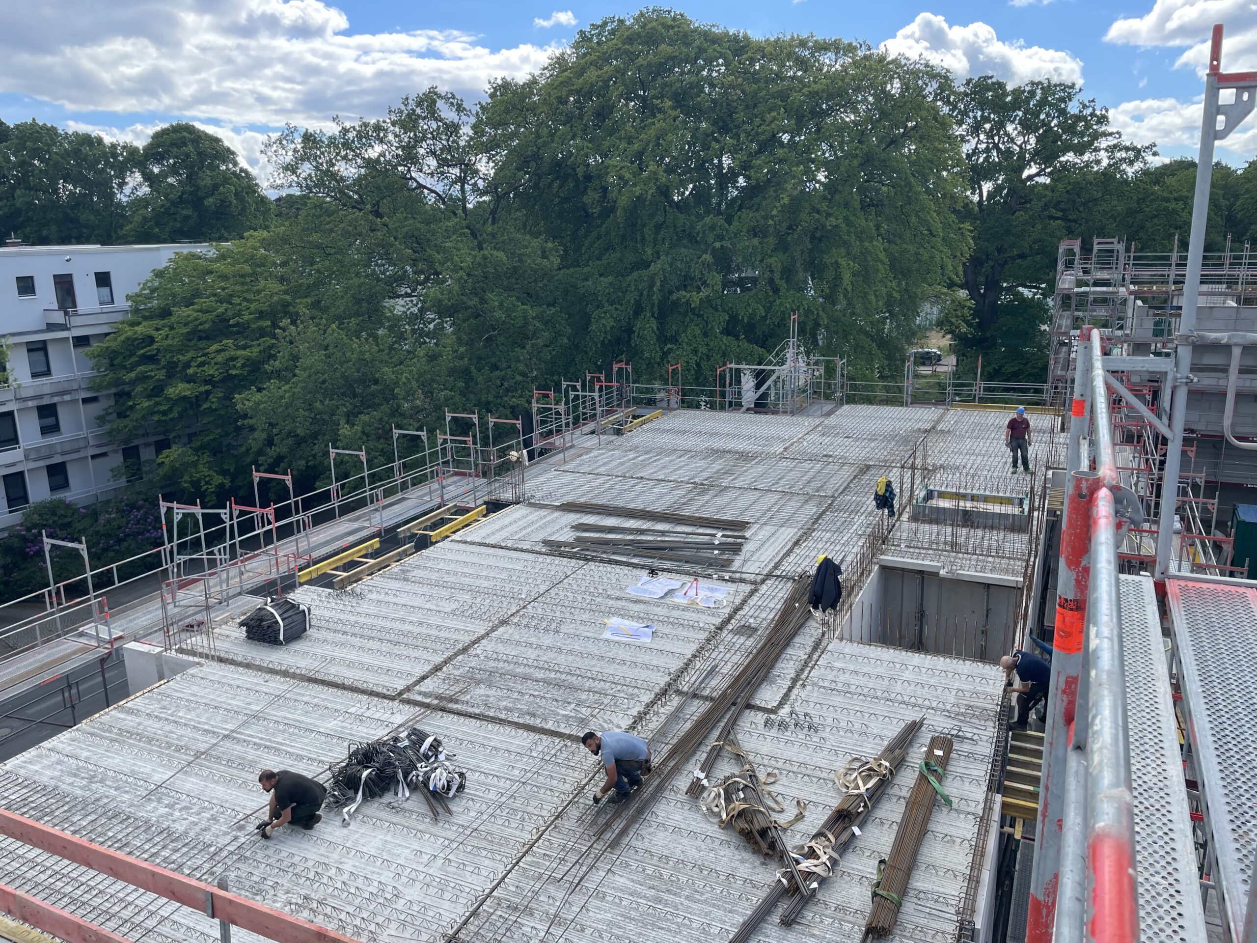 Bauarbeiter arbeiten auf einer Baustelle mit Stahlträgern und Bäumen im Hintergrund.