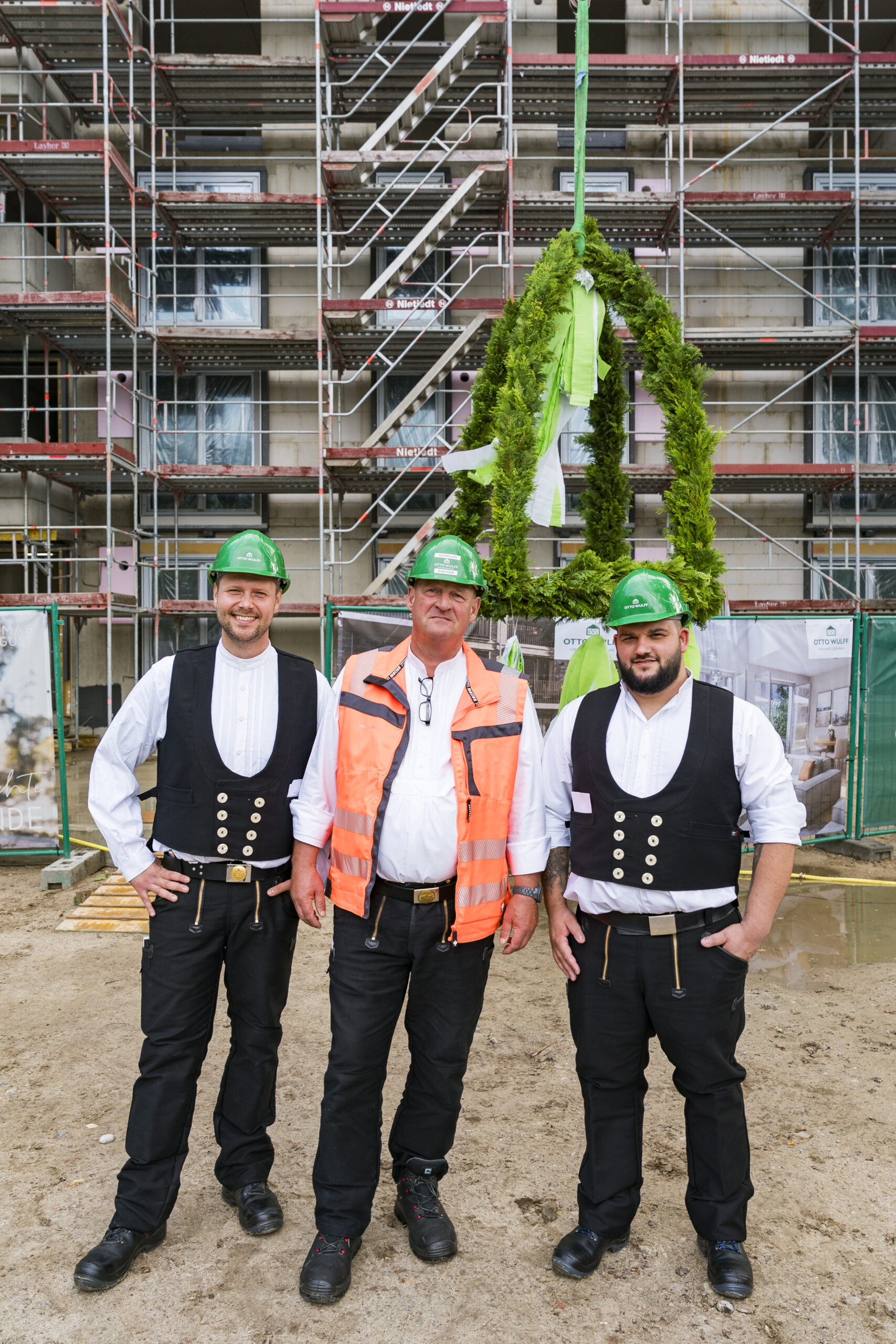 Drei Bauarbeiter vor Richtkranz und Gerüst, tragen grüne Helme und Zunftkleidung.