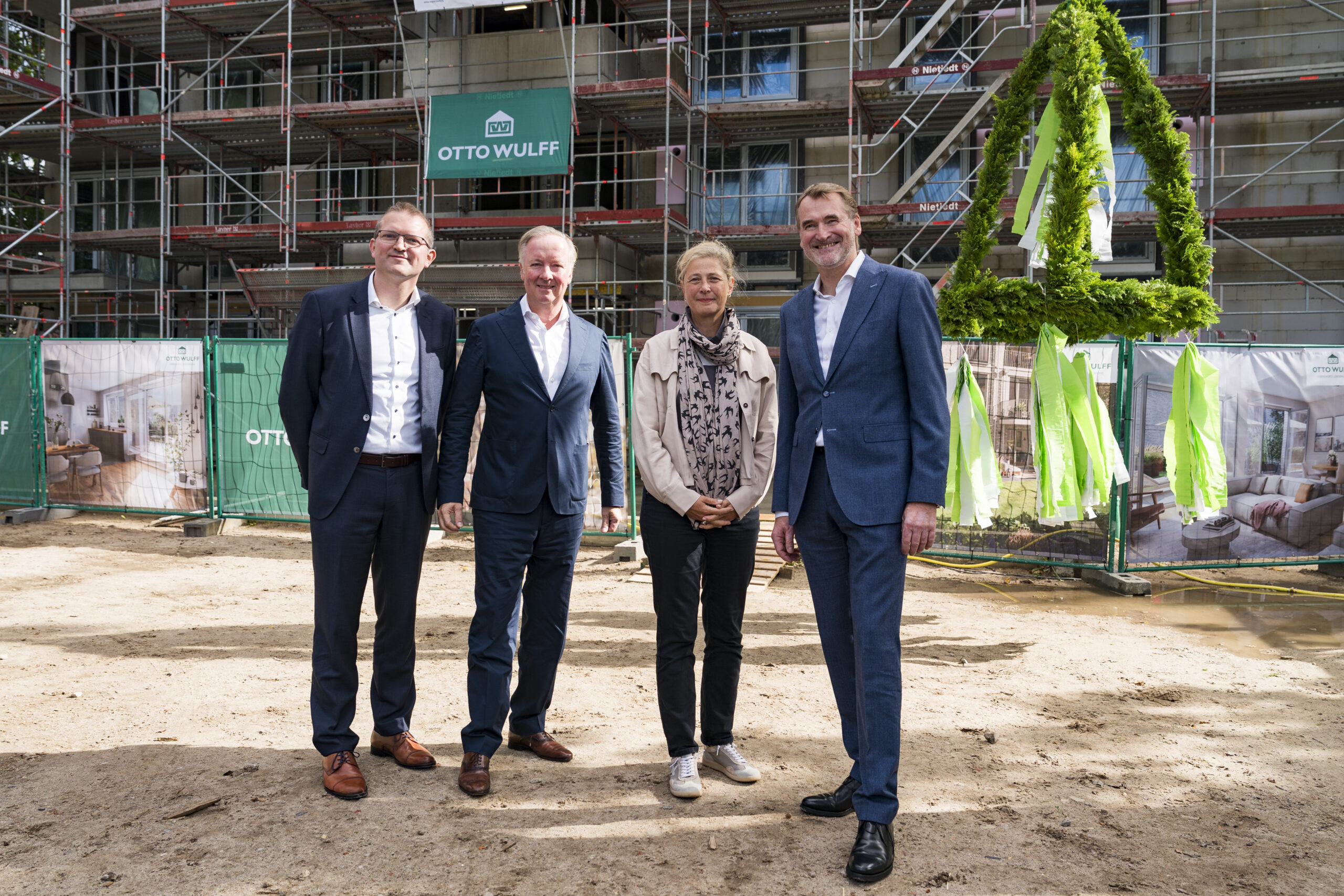 Vier Personen stehen vor einer Baustelle mit Richtkranz und grünen Absperrungen.