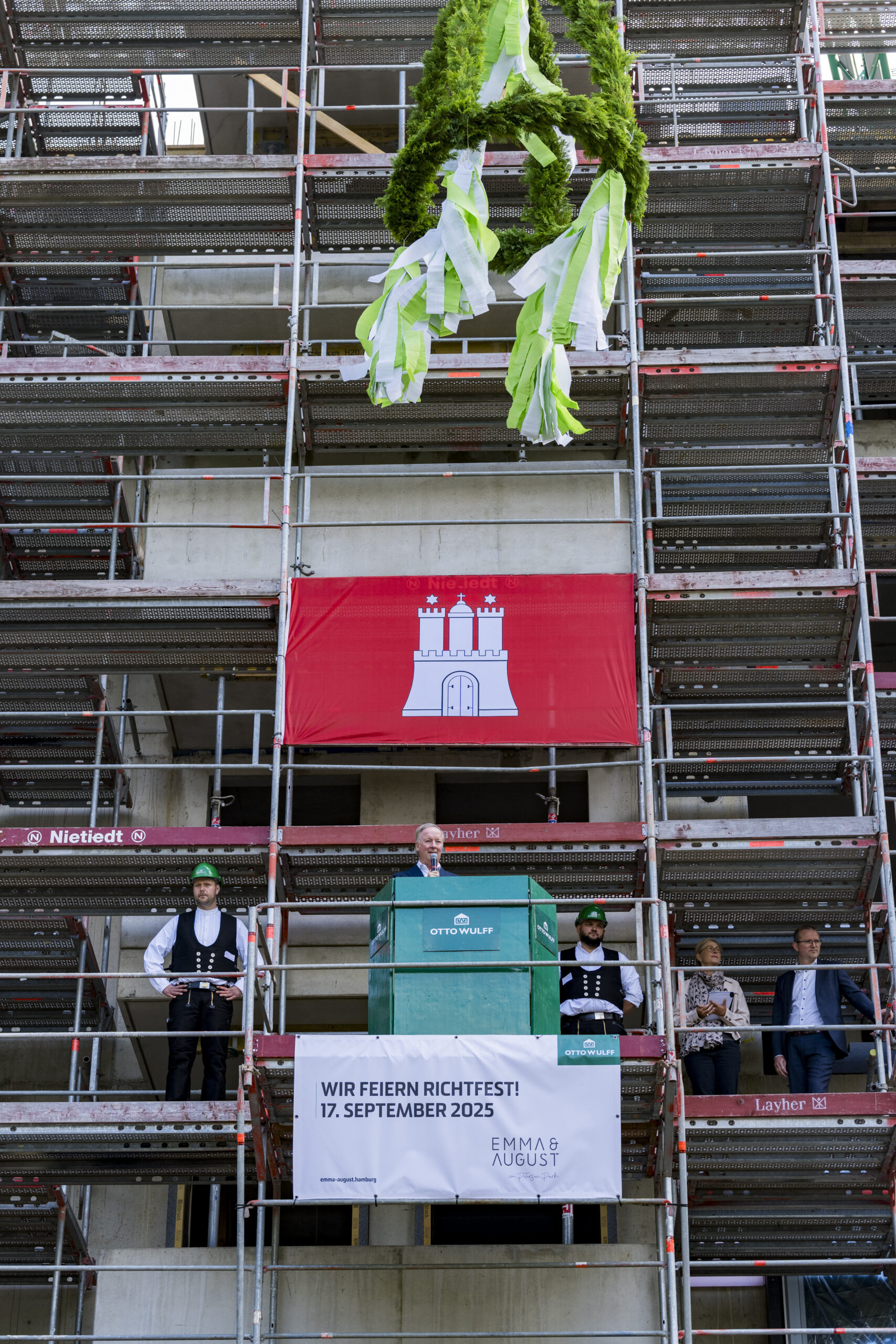 Richtfest an Baustelle mit Redner, Kranz und Hamburg-Flagge im Hintergrund.