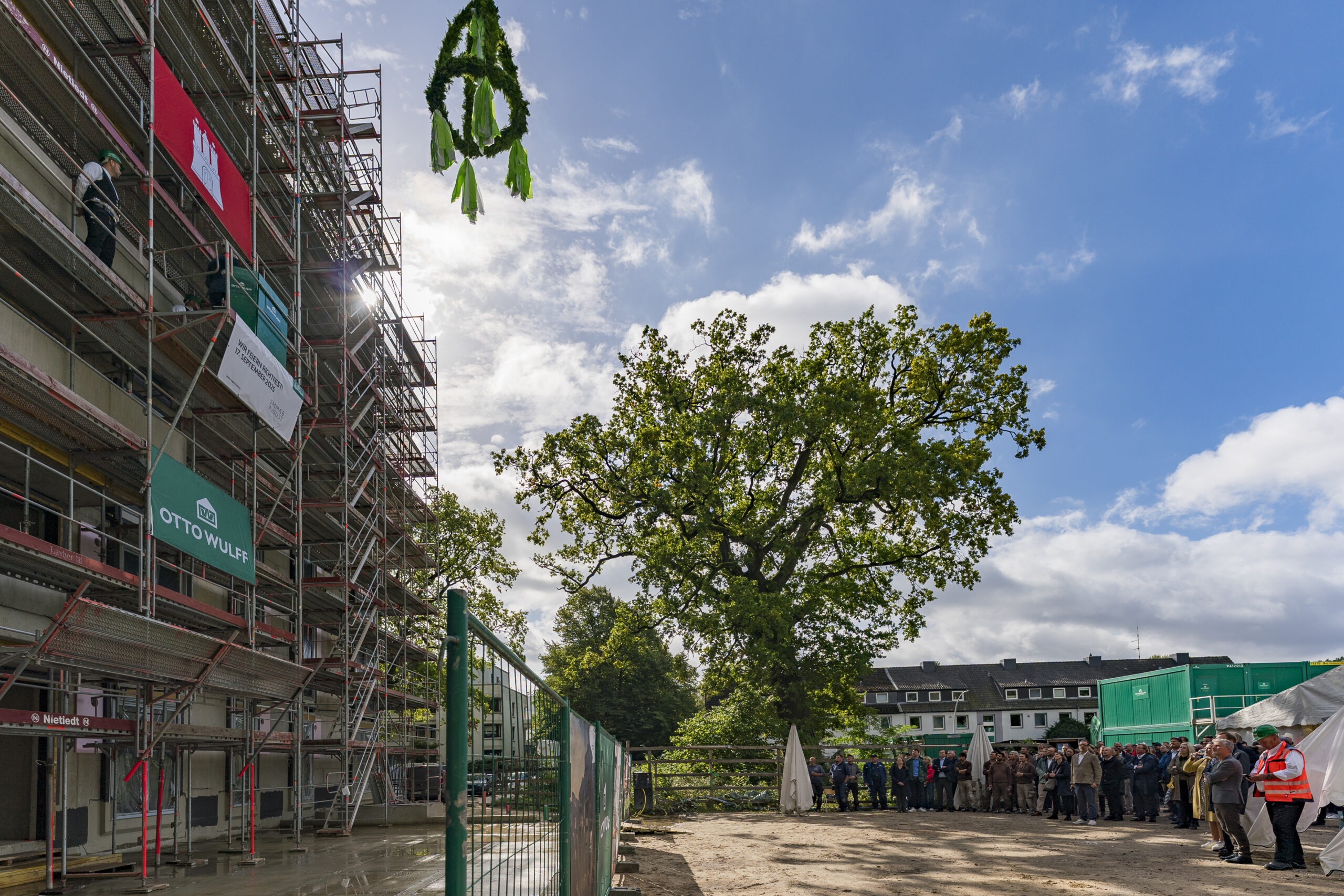 Richtfest an einer Baustelle mit Baugerüst, Baum, Publikum und Richtkranz.