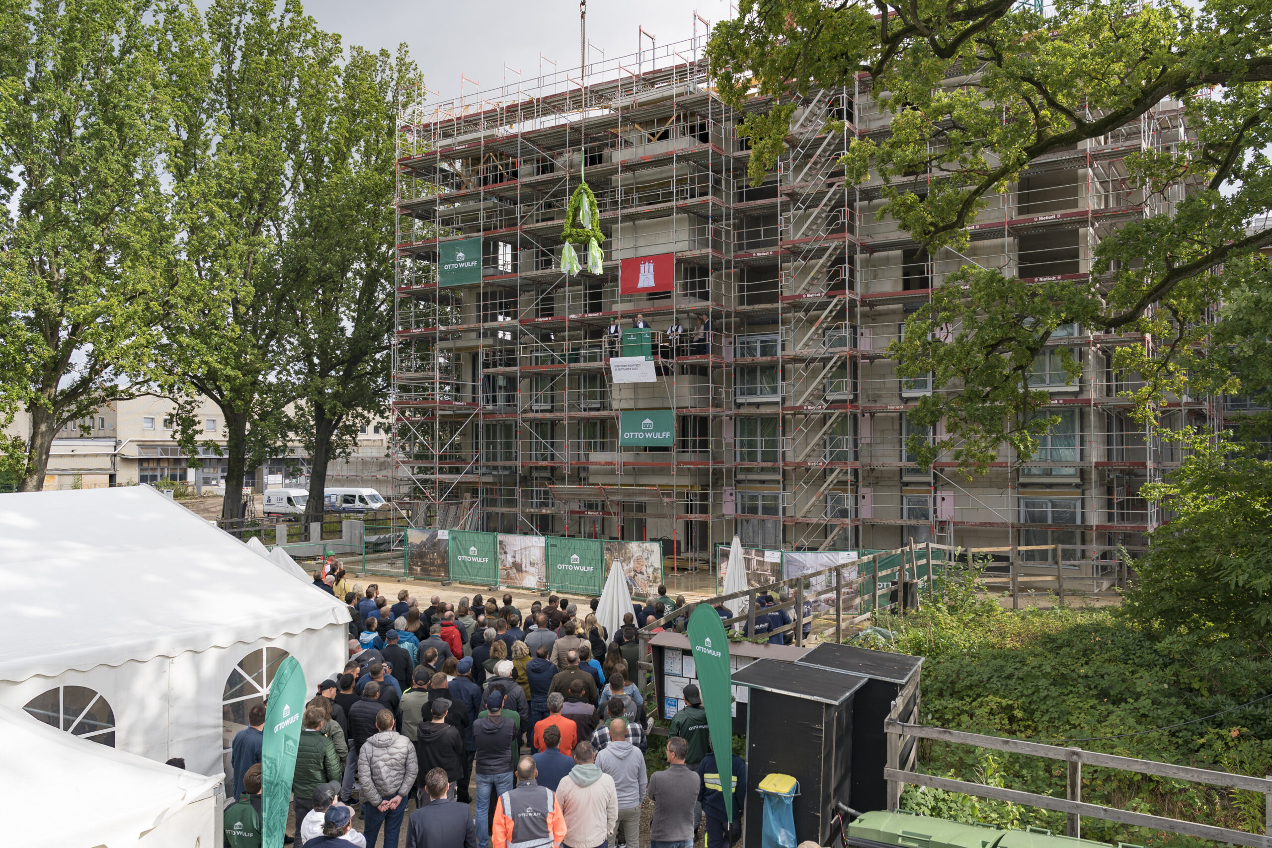 Menschenmenge vor Rohbau mit Richtkranz, Baugerüst und großen Bäumen.