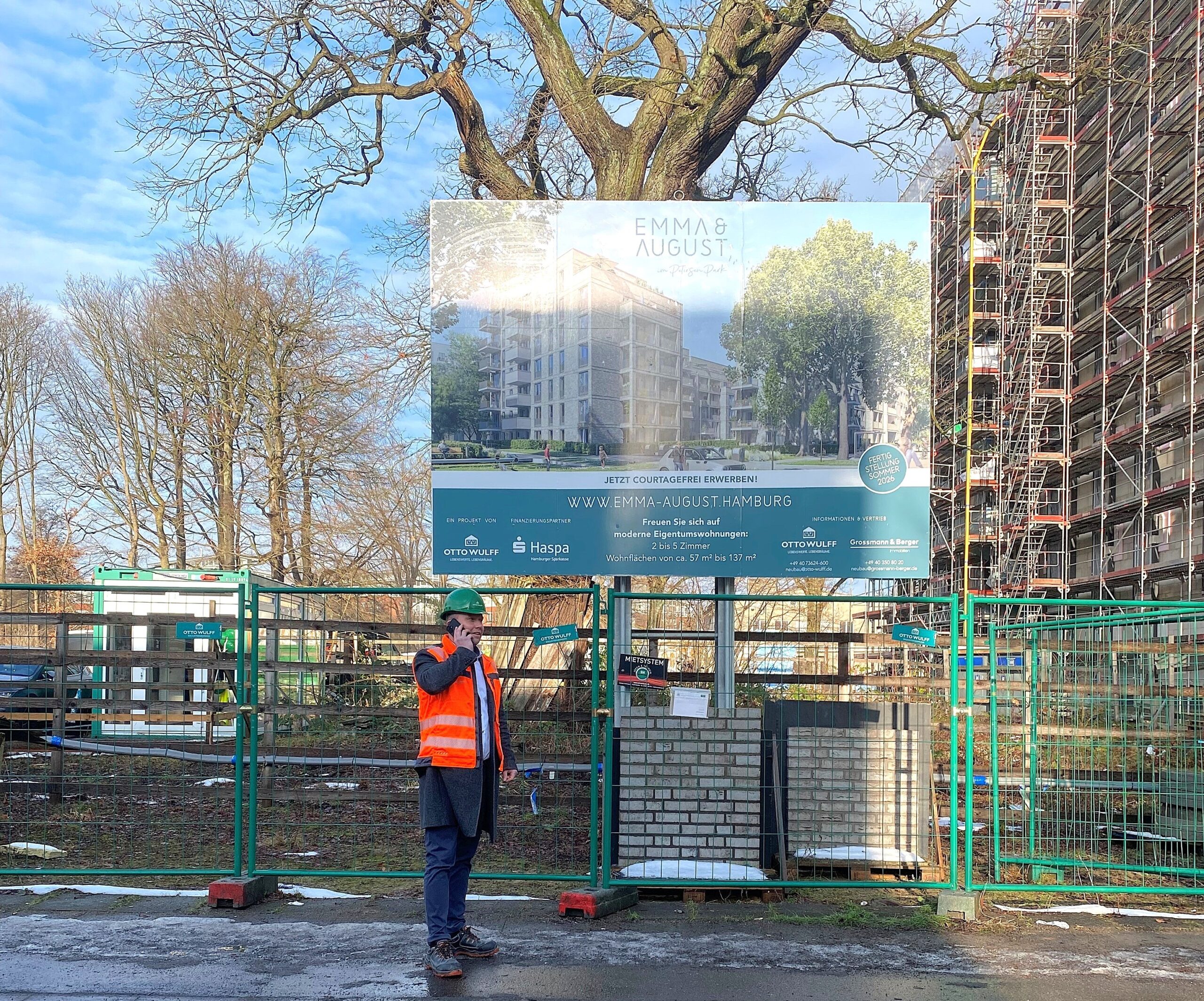 Bauarbeiter mit Weste und Helm telefoniert vor Bauschild und Baustelle.