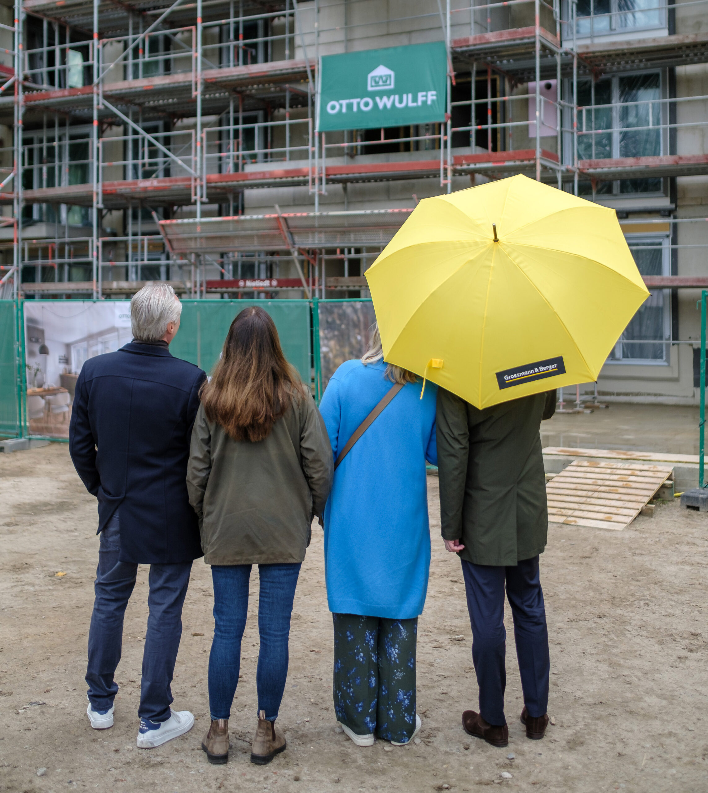 Vier Personen stehen vor einer Baustelle, eine hält einen großen gelben Regenschirm.