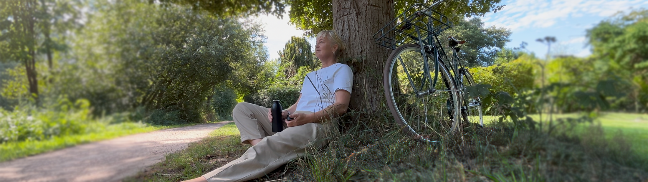 Header 1 Person sitzt am Baum mit Fahrrad und entspannt in grüner Natur an einem Weg.